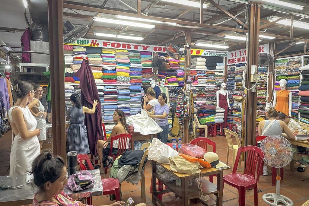 Hoi An Central Market tailor shop – bustling fabric stalls with international visitors getting fitted for custom clothes, showcasing Hoi An’s famous tailoring culture.