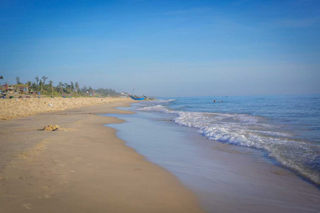 Wide sandy shoreline of Tam Thanh Beach in Quang Nam, Vietnam, with gentle waves rolling in under a clear blue sky.