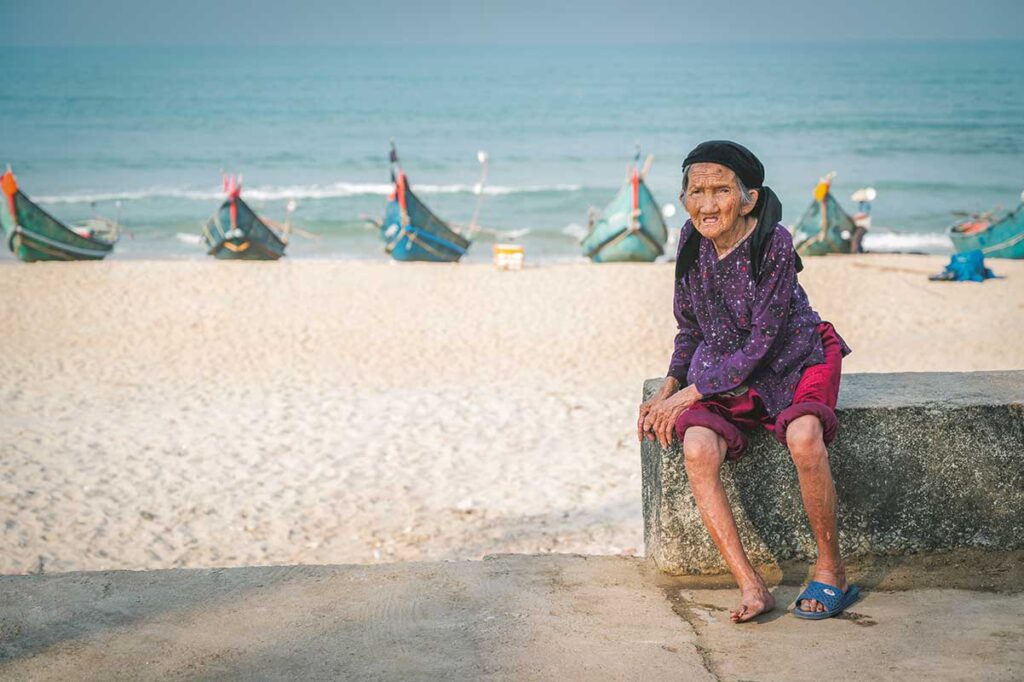 Elderly woman sitting by Tam Thanh Beach with colorful fishing boats lined up on the sand, reflecting the charm of Vietnam’s fishing villages.