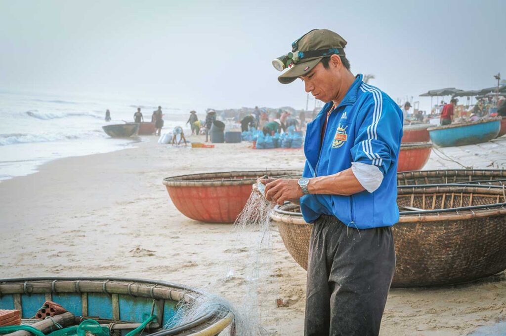 Vietnamese fisherman repairing his fishing net beside a coracle boat at Tam Tien Beach.