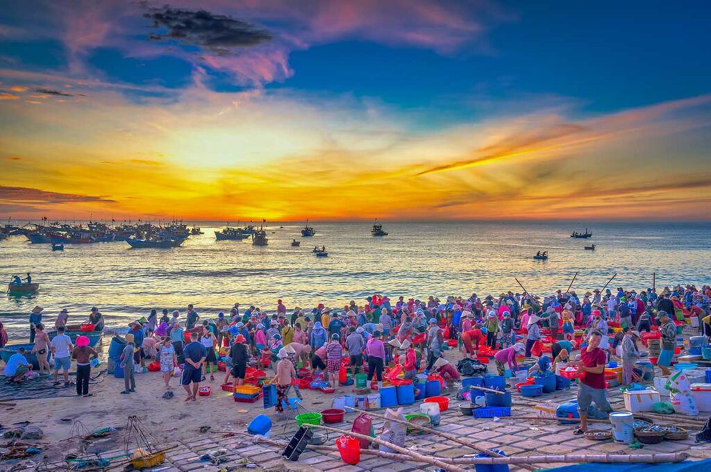 Crowded Tam Tien beach fish market at dawn with colorful buckets and fishing nets.
