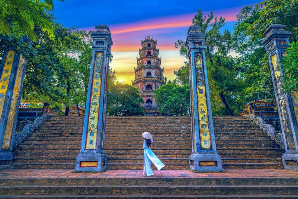 A Vietnamese woman in Ao Dau and conical gate posing near the stairs leading to the main stupa tower at Thien Mu Pagoda in Hue