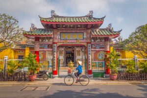 Cyclist passing Trieu Chau Assembly Hall in Hoi An Ancient Town – Daily life scene with a local woman cycling past the colorful Chinese-style temple gate.