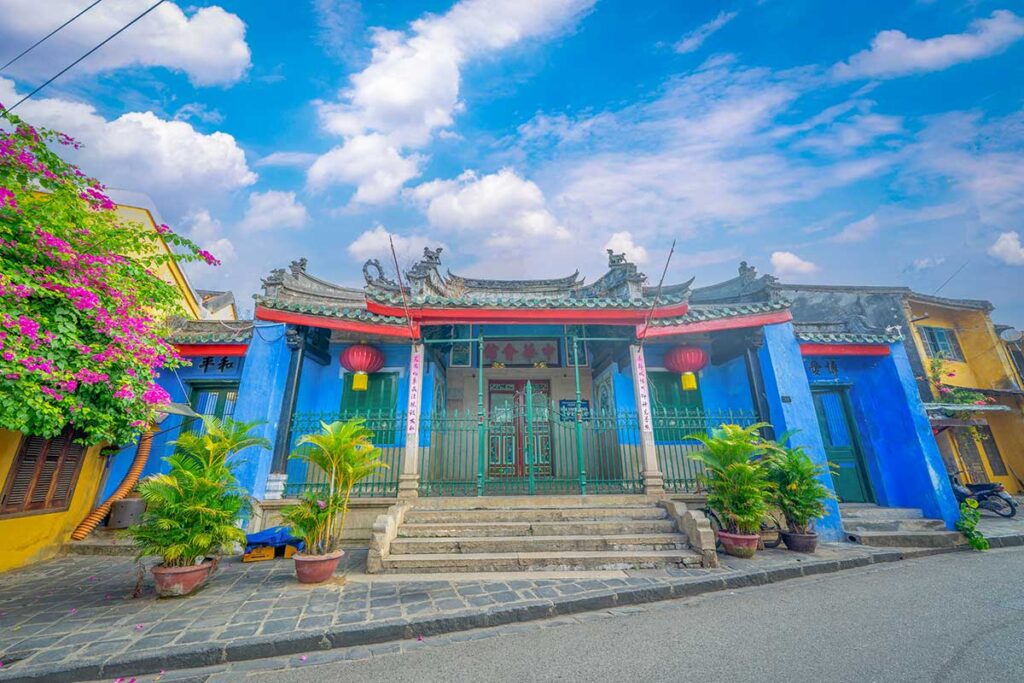 Blue-painted facade of Trung Hoa Assembly Hall in Hoi An, with red lanterns, green iron railings, and Chinese calligraphy.