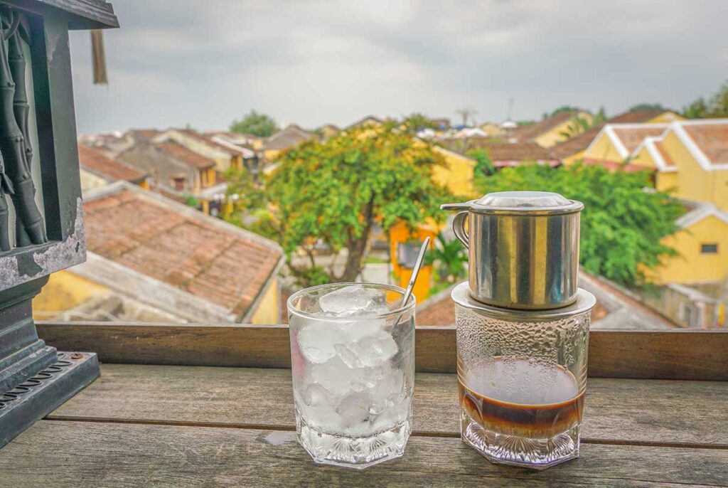 Traditional Vietnamese iced coffee served with a phin filter overlooking the yellow-tiled rooftops of Hoi An Ancient Town from a local café balcony.