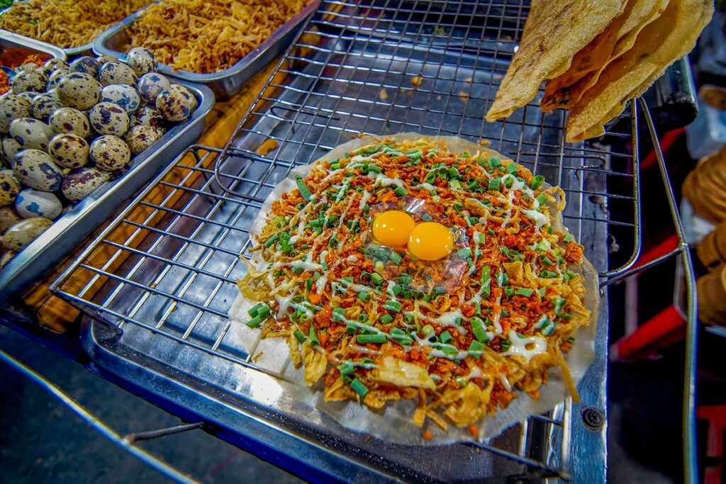Crispy Vietnamese pizza (bánh tráng nướng) topped with quail eggs, scallions, and dried toppings grilling on a street food stall in Hoi An.