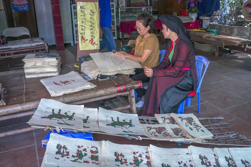 In the foreground a few Dong Ho Painting and in the background two woman making them at Kids are sitting on a mat with a man in traditional Vietnamese clothes and showing the children how to make a traditional Dong Ho Painting during a workshop in Dong Ho Painting Village