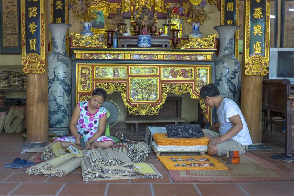 A man and a woman sitting in front of a family altar in a house working on Dong Ho Paintings at Dong Ho Painting Village