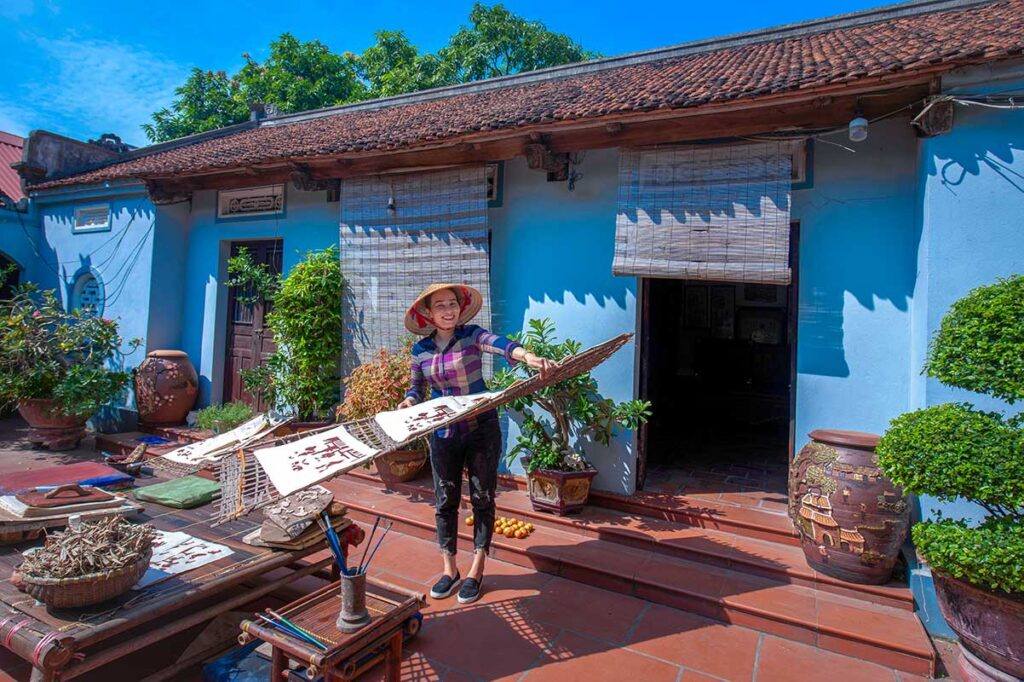 A woman standing in front of a local house and holden a bamboo drying rack with Dong Ho paintings at Kids are sitting on a mat with a man in traditional Vietnamese clothes and showing the children how to make a traditional Dong Ho Painting during a workshop in Dong Ho Painting Village
