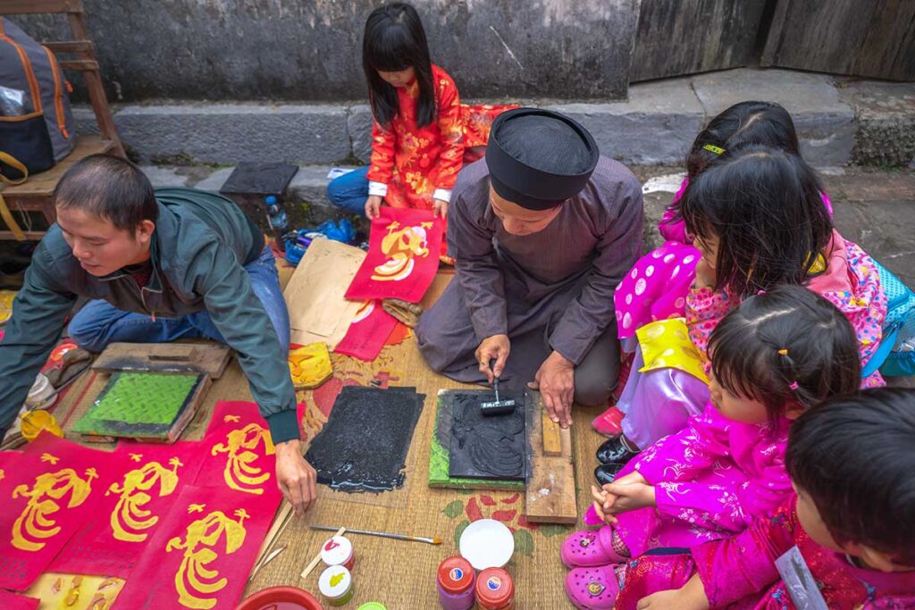 Kids are sitting on a mat with a man in traditional Vietnamese clothes and showing the children how to make a traditional Dong Ho Painting during a workshop in Dong Ho Painting Village