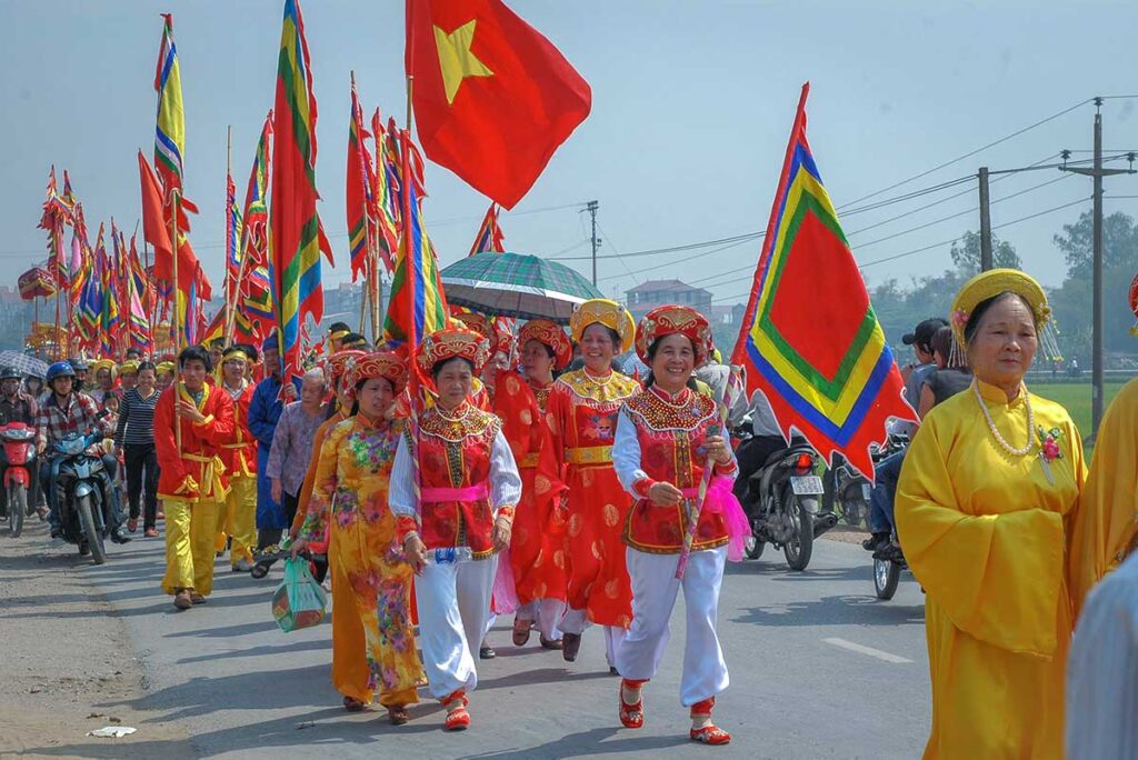 A parade with flags and people with colorful traditional clothes, mainly red and yellow walking through a street during Lim Festival in Bac Ninh 