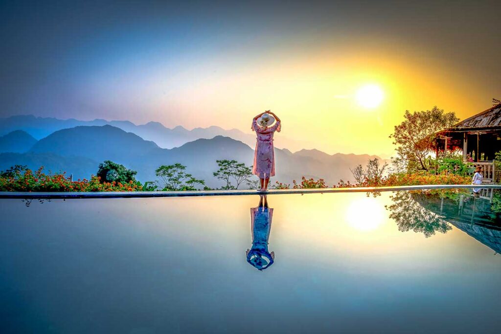 A woman standing on the edge of an infinity pool and a luxury lodge at sunset with blue sky and beautiful weather in Pu Luong