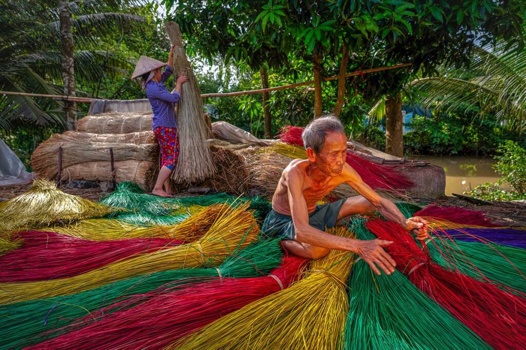 Artisans preparing dyed sedge in bright colors of red, yellow, and green along a canal at Dinh Yen Mat Weaving Village