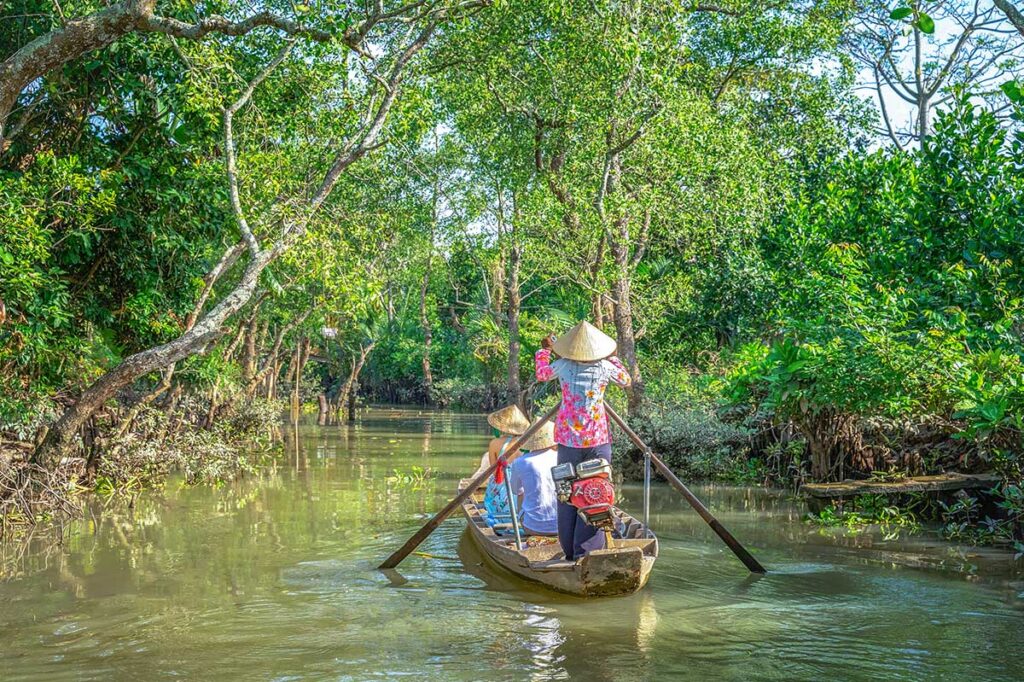 Tourists on a traditional sampan boat paddling through shaded canals in Tien Giang, a classic Mekong Delta experience.