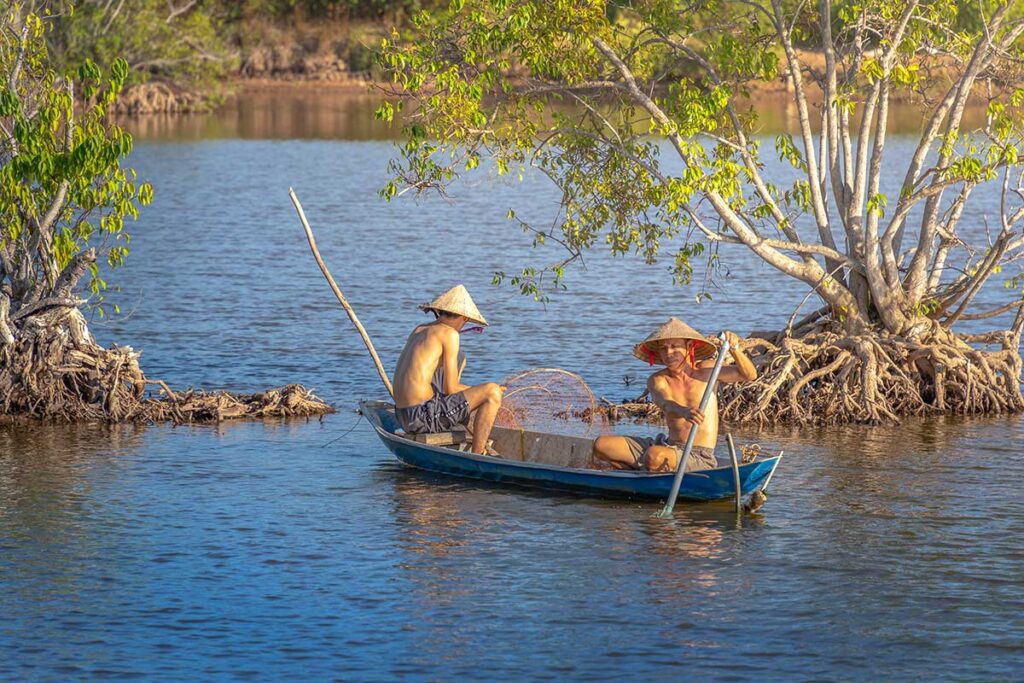 Two shirtless Vietnamese fishermen navigate a small wooden boat through Tra Vinh’s flooded mangrove forest, checking handmade fish traps in calm waters.