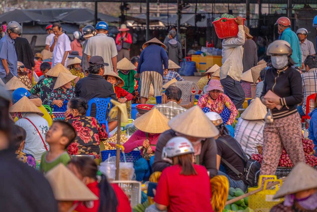 Crowded scene at Vi Thanh Market with women in conical hats selling potatoes, bananas, and vegetables