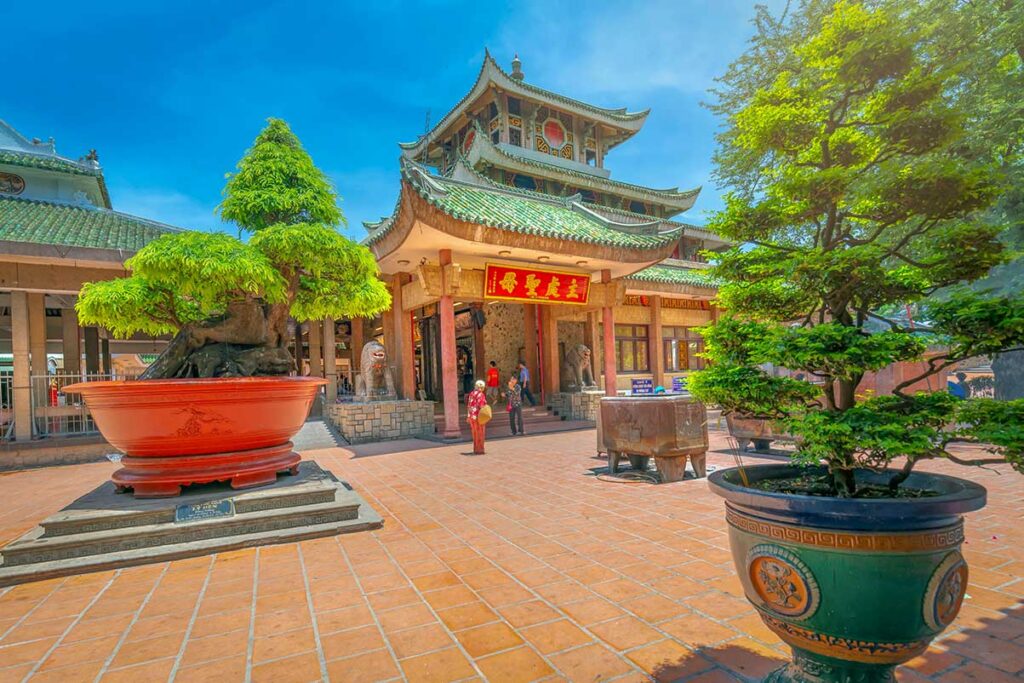 Front courtyard of Ba Chua Xu Temple in Chau Doc, An Giang, with bonsai trees and the multi-tiered roofed entrance on a sunny day.