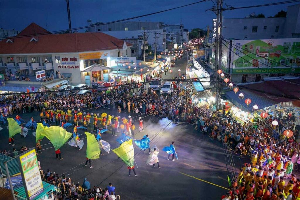 Street parade during Ba Chua Xu Festival in Chau Doc, An Giang, Vietnam, with colorful flags, lion dance troupes, and large crowds lining the streets at night.