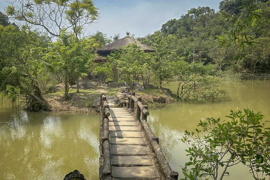 Stone bridge leading to a thatched pavilion at Huyen Khong Son Thuong Pagoda, surrounded by lush trees and tranquil water in Hue’s forested hills.