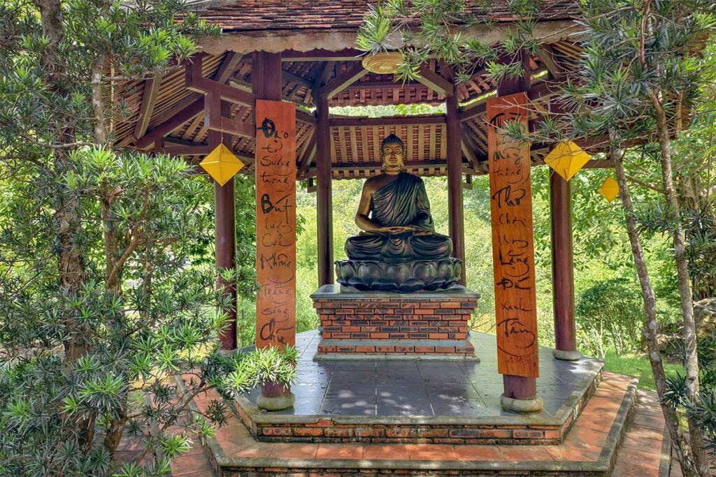 Outdoor Buddha statue in a shaded wooden pavilion, decorated with calligraphy pillars at Huyen Khong Son Thuong Monastery near Hue.