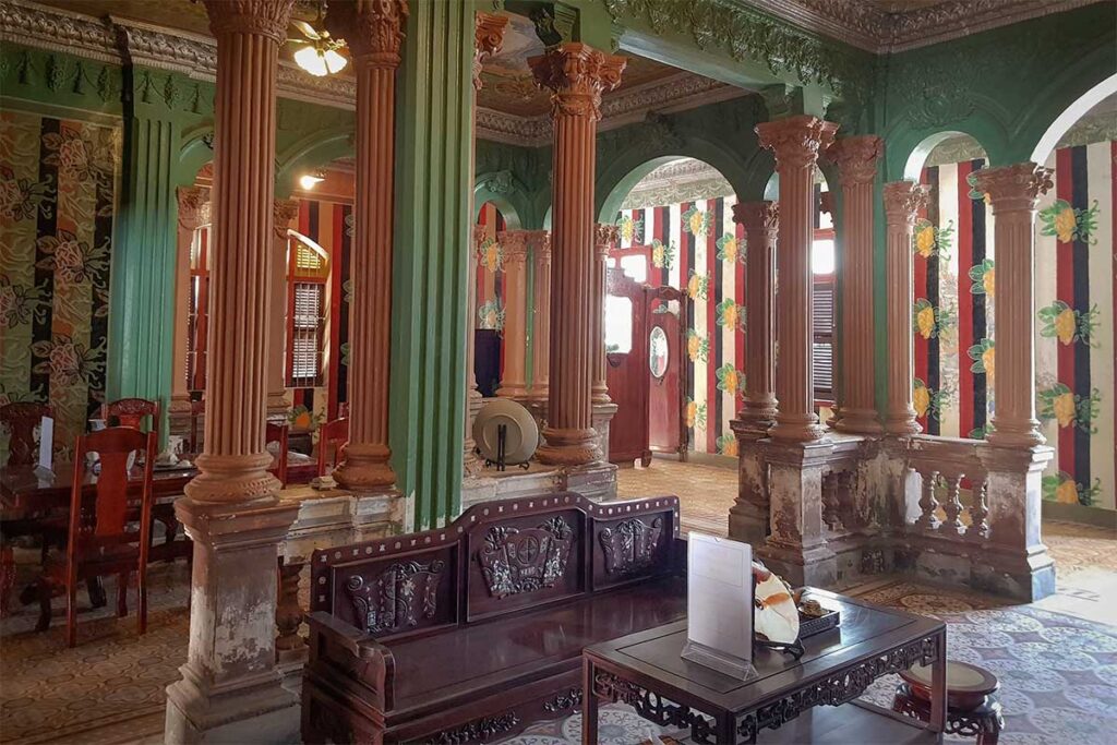 Interior of Huynh Ky Ancient House featuring European-style columns, antique wooden furniture, and patterned walls.