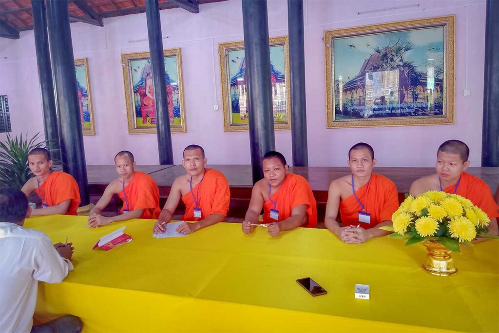 Khmer monks in orange robes sitting at a long yellow table inside Nodol Pagoda, Tra Vinh