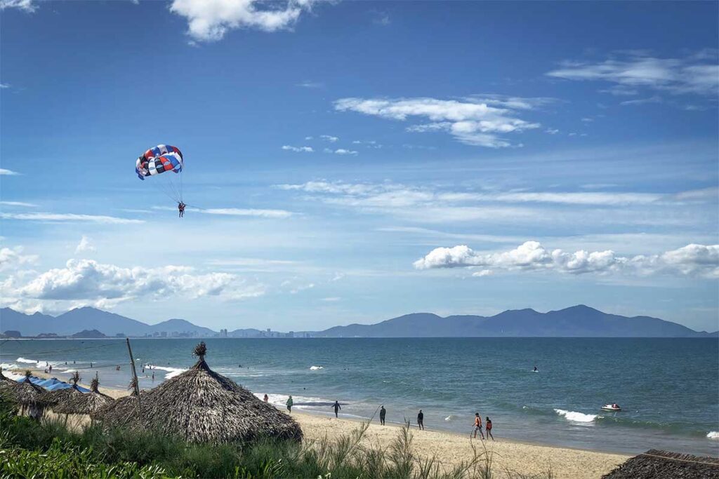 Parasailing and beach activities at An Bang Beach Hoi An, with views of the sea, palm huts, and Da Nang mountains in the distance.