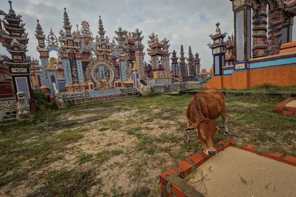 Colorful mausoleums at An Bang Cemetery in Hue, Vietnam, rise above sandy ground while a cow grazes nearby — a striking mix of rural life and ornate ancestral tombs.