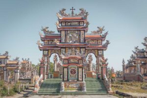 Front view of an elaborate family tomb at An Bang Cemetery near Hue, with dragon statues, ceramic mosaics, and multi-tiered roofs gleaming under the sun.