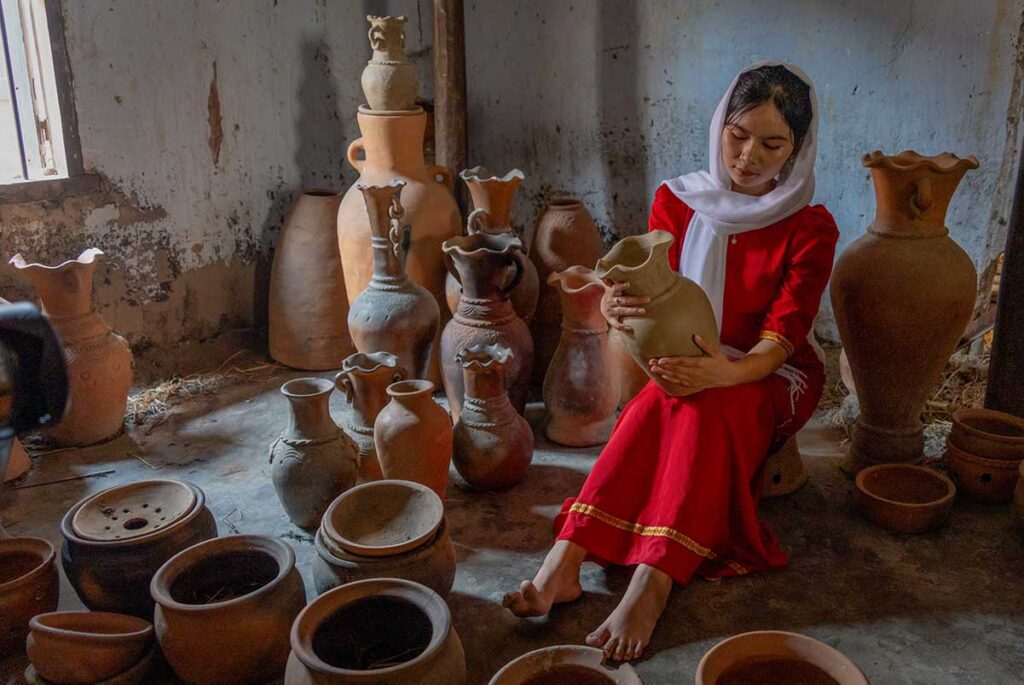 Cham woman in traditional dress holding handmade pottery inside Bau Truc Pottery Village, surrounded by terracotta vases and jars.