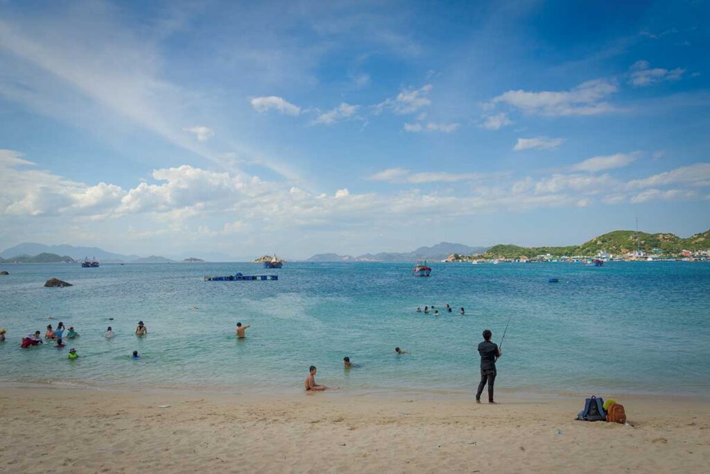 Binh Hung Beach at Vinh Hy Bay with swimmers and fishing boats under blue sky – A lively scene of locals swimming, fishing, and relaxing on the sandy shore with fishing boats anchored in the turquoise waters of Vinh Hy Bay, Ninh Thuan.