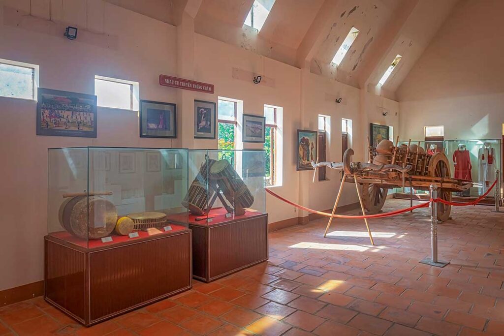 Exhibition inside the Cham Ethnic Museum at Po Klong Garai complex, displaying traditional musical instruments, costumes, and a wooden buffalo cart.