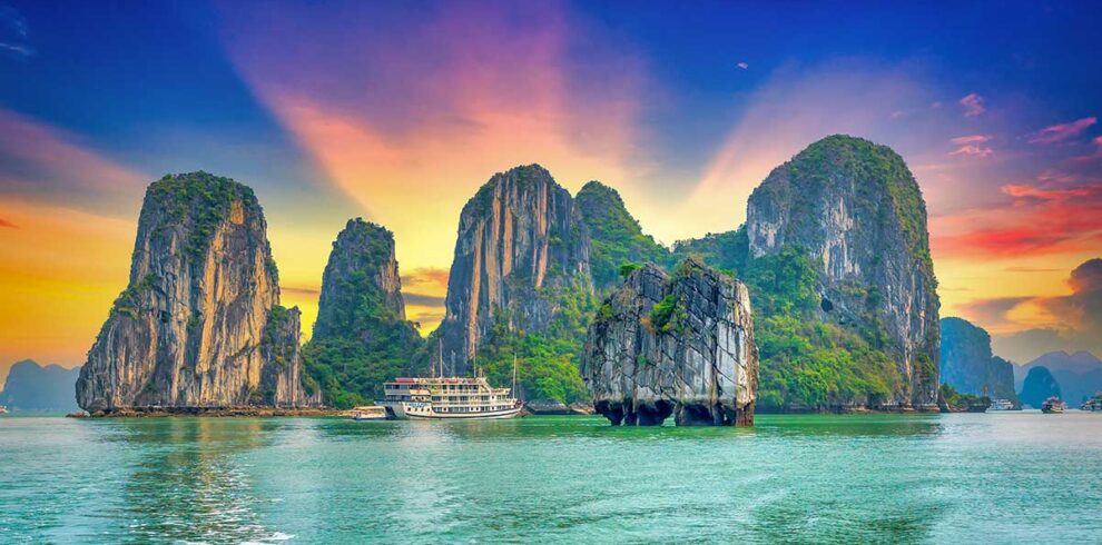 Halong Bay tours – Local Vietnam | cruise boat sailing between limestone karsts at sunset on Vietnam’s UNESCO World Heritage bay