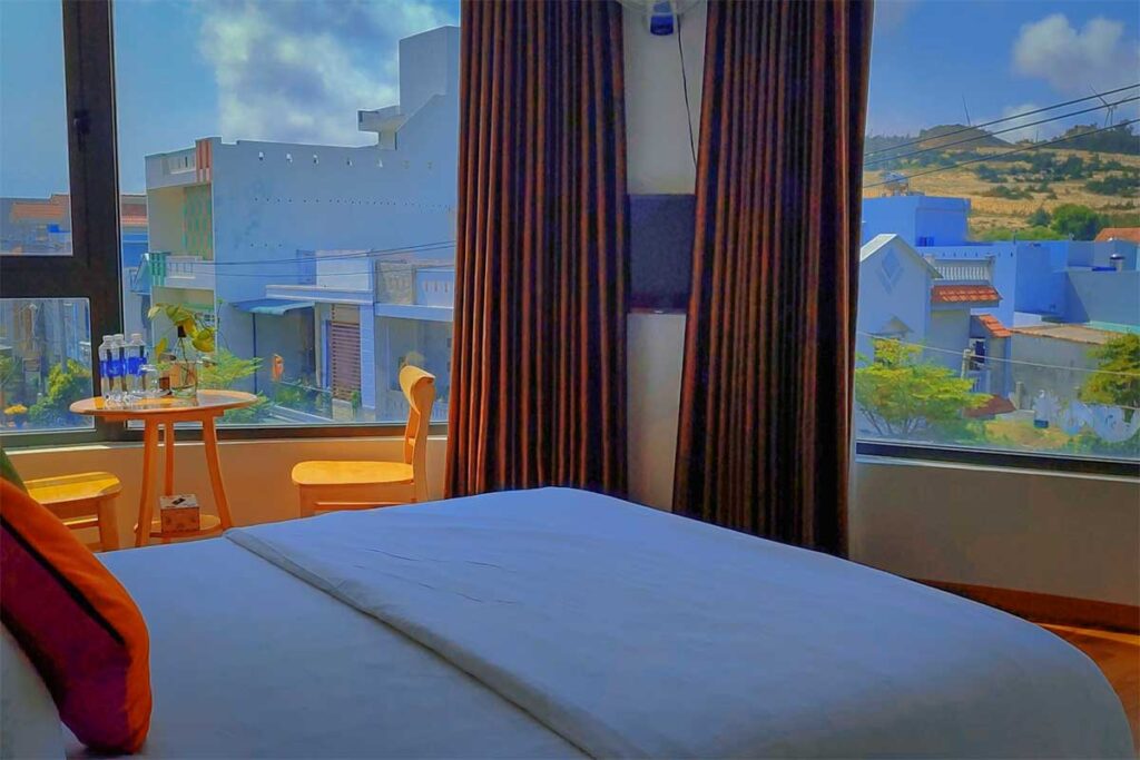 Simple homestay room in Nhon Ly fishing village with window views over blue-and-white houses and hills—budget base for trips to Ky Co and Eo Gio.