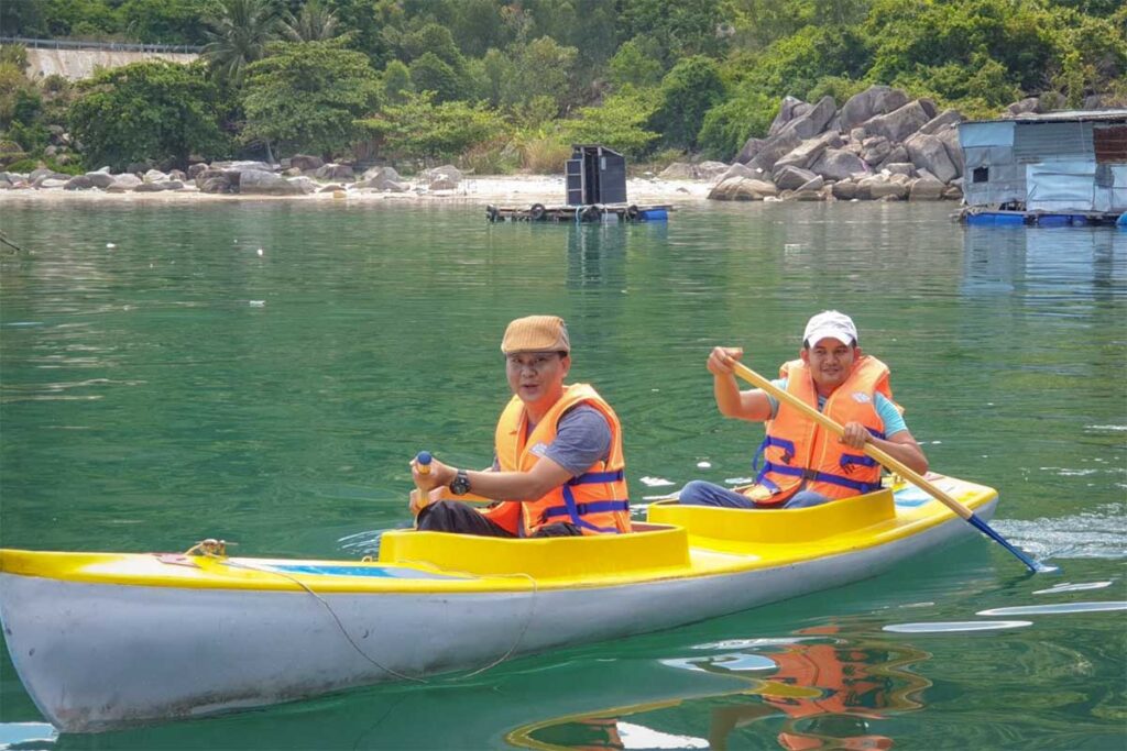 Two men kayaking in the calm emerald waters of Vung Ro Bay, with floating houses and forested hills in the background.