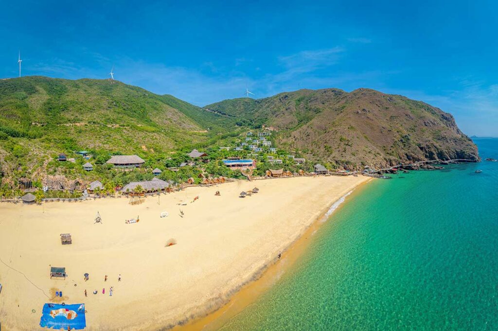Aerial panorama of Ky Co Beach, Quy Nhon, Vietnam: broad golden sand, emerald water, cliff-backed bay with rustic huts and wind turbines on the ridge—a signature stop among the best beaches in Quy Nhon.