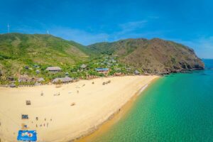 Aerial panorama of Ky Co Beach, Quy Nhon, Vietnam: broad golden sand, emerald water, cliff-backed bay with rustic huts and wind turbines on the ridge—a signature stop among the best beaches in Quy Nhon.