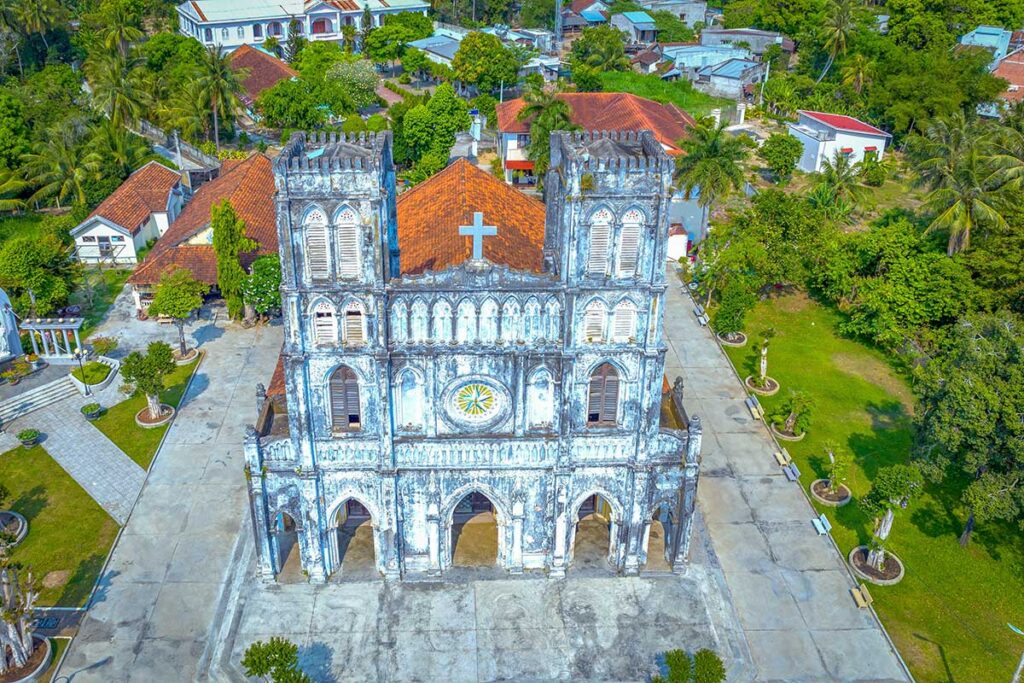 Frontal aerial shot of Mang Lang Church with twin bell towers, faded Gothic carvings, and open courtyard.