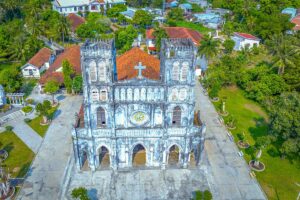Frontal aerial shot of Mang Lang Church with twin bell towers, faded Gothic carvings, and open courtyard.