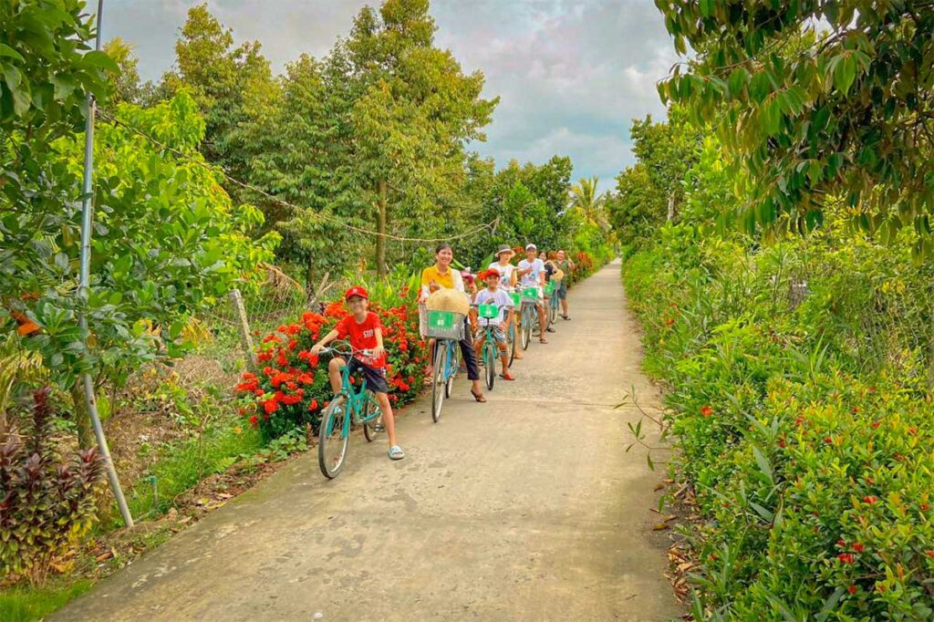 Family cycling with children along a quiet countryside path in the Mekong Delta, surrounded by tropical greenery and orchards.