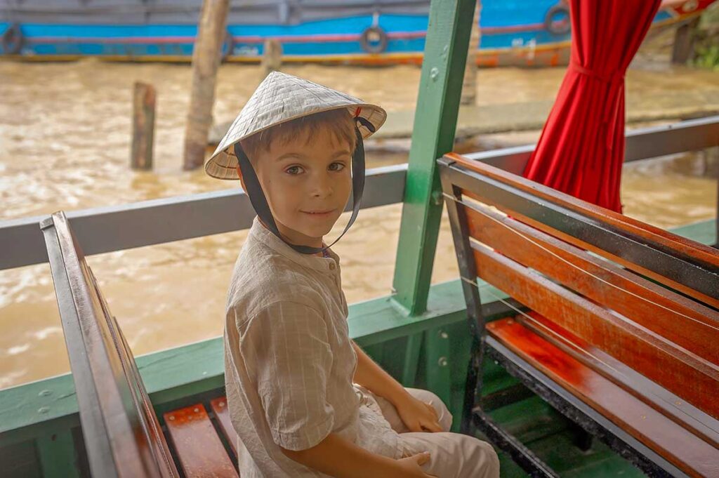 Young boy in a Vietnamese conical hat enjoying a boat ride through the Mekong Delta, a family-friendly way to explore the region with kids.