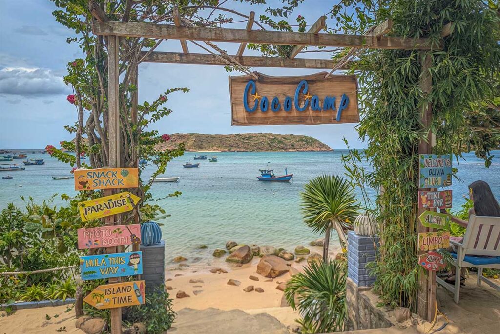 CocoCamp in Nhon Hai – A wooden sign leading down to the beach, with basket boats floating in turquoise water and Hon Kho Island across the bay.