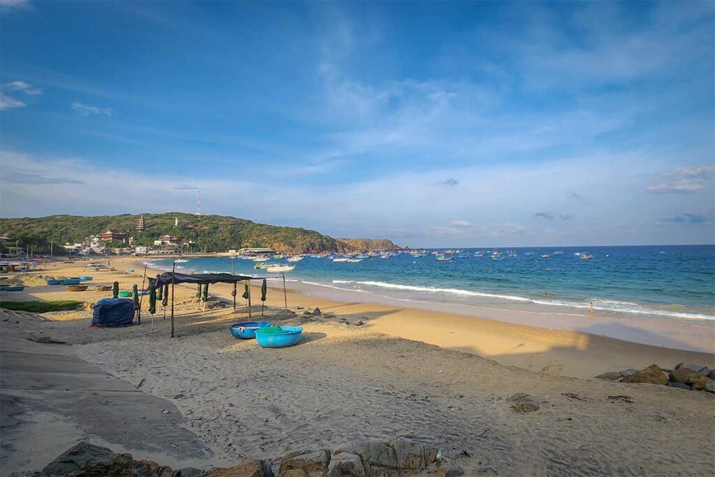 Wide sandy arc of Nhon Ly Beach with calm blue water, fishing boats and headland views near Quy Nhon.