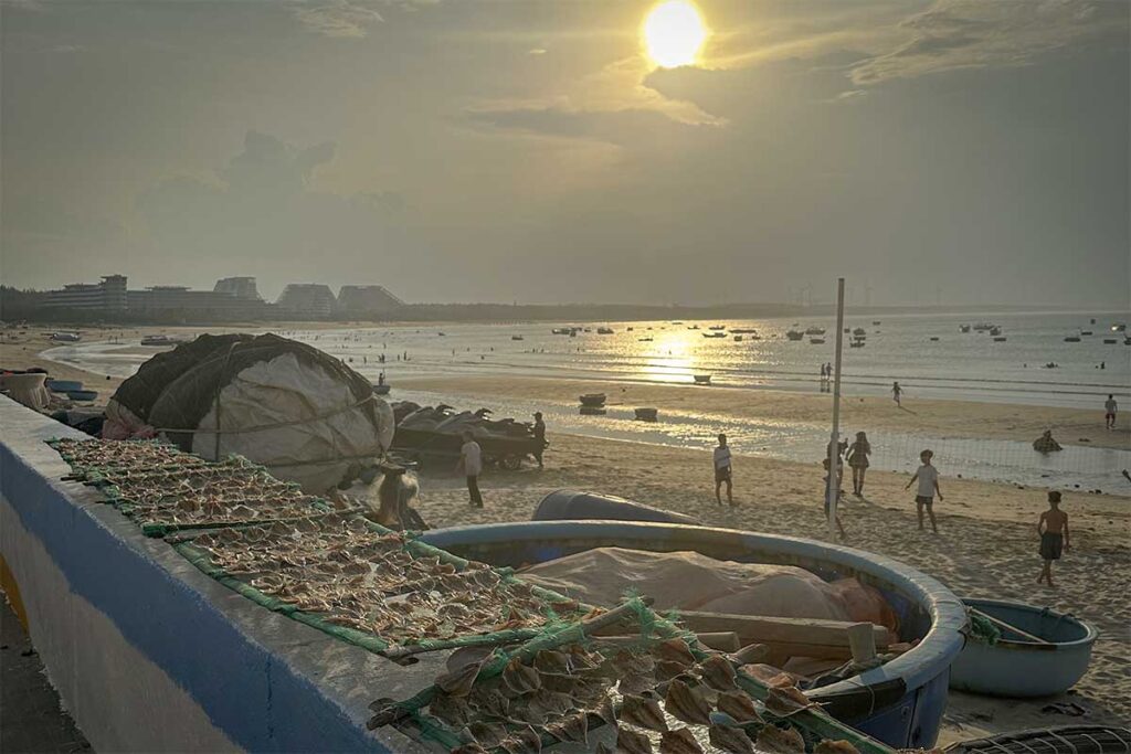 Sunset on Nhon Ly Beach, Quy Nhon: locals playing on wide tidal flats while fishing boats rest offshore and dried seafood racks line the seawall.