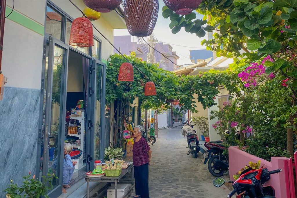 Lantern-strung alley in Nhon Ly fishing village, Quy Nhon with a tiny street stall, bougainvillea, and motorbikes—everyday local life steps from the beach.