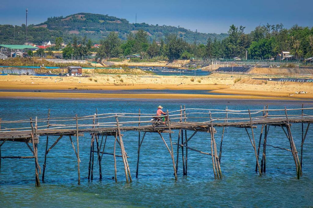 Local riding a motorbike across Ong Cop Wooden Bridge in Phu Yen, Vietnam, with sandy banks and aquaculture farms in the background.