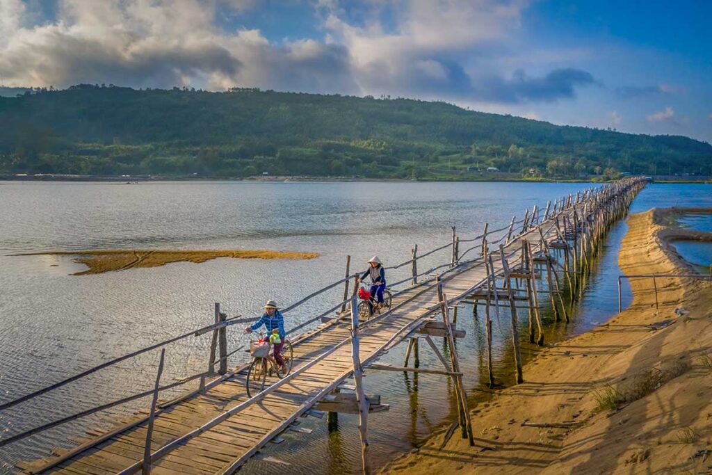 Two women in conical hats cycling across Ong Cop Wooden Bridge, with sandy riverbanks and green hills in the distance.