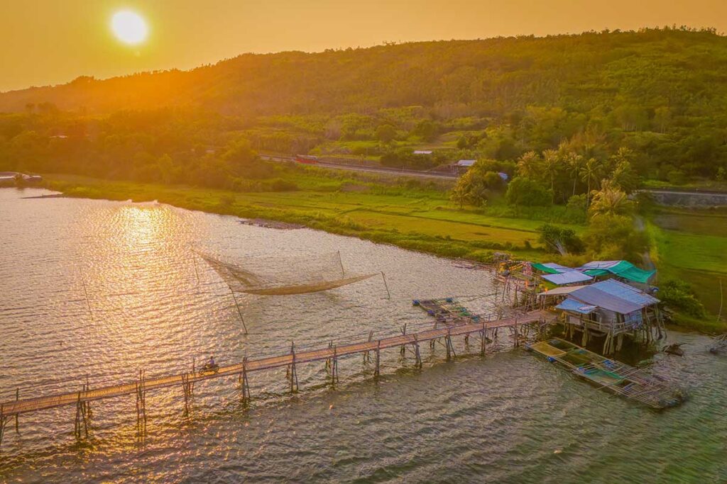 Aerial sunset view of Ong Cop Wooden Bridge and riverside houses in Phu Yen, with fishing nets spread across the water.