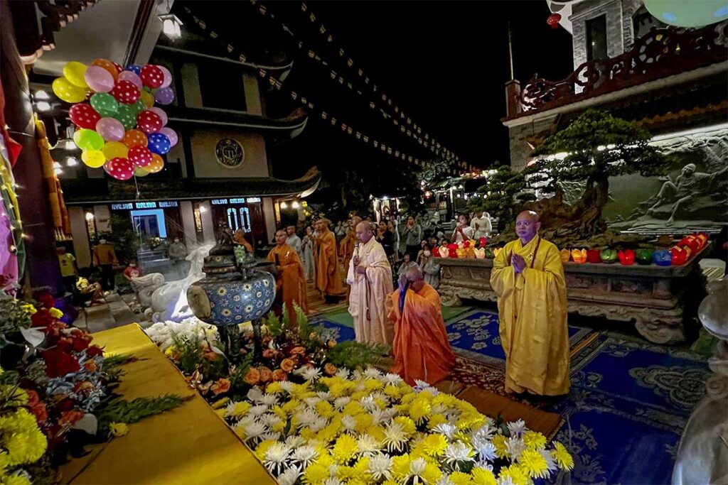 Evening ceremony at Thien Hung Pagoda with monks in saffron robes, flowers, incense, and worshippers gathered in the courtyard.