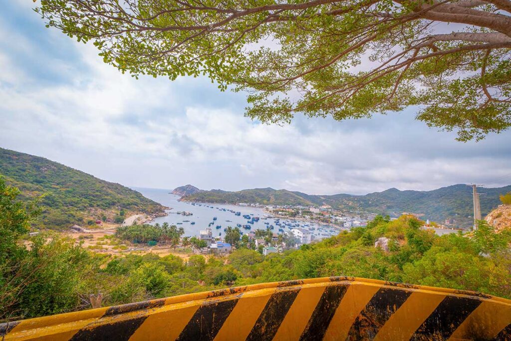 Scenic overlook of Vinh Hy Bay, framed by tree branches, showing the fishing harbor and coastal village inside Nui Chua National Park.